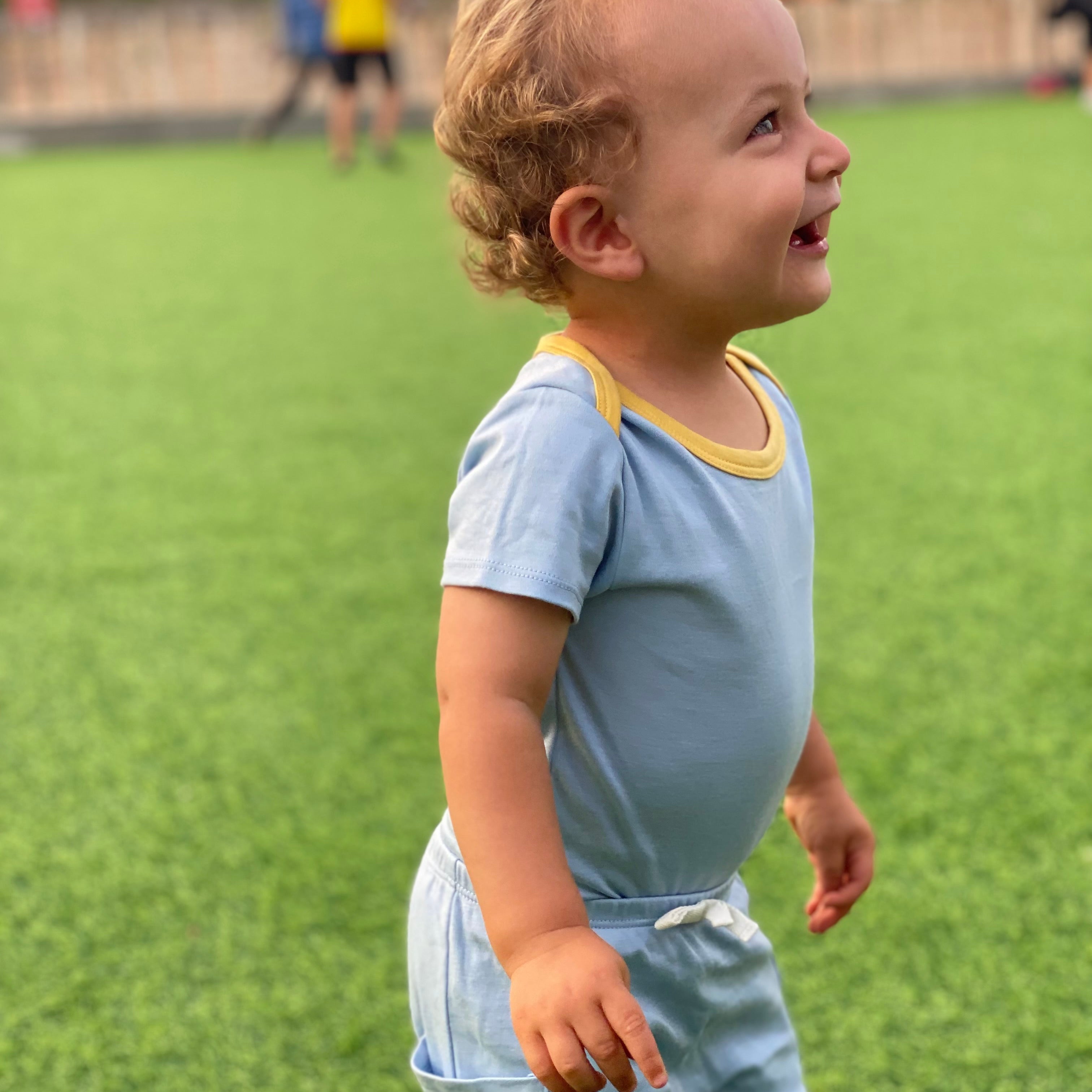 Chambray Blue with Yellow | Child in light blue outfit standing on a grassy field with other people in the background