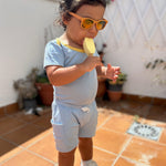 Chambray Blue with Yellow | Child wearing sunglasses, a sunny day set with shorts and short sleeves onesie and eating a popsicle on a tiled patio