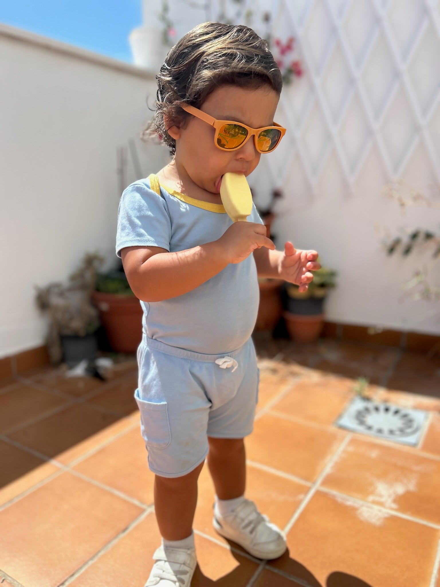 Chambray Blue with Yellow | Child wearing sunglasses, a sunny day set with shorts and short sleeves onesie and eating a popsicle on a tiled patio