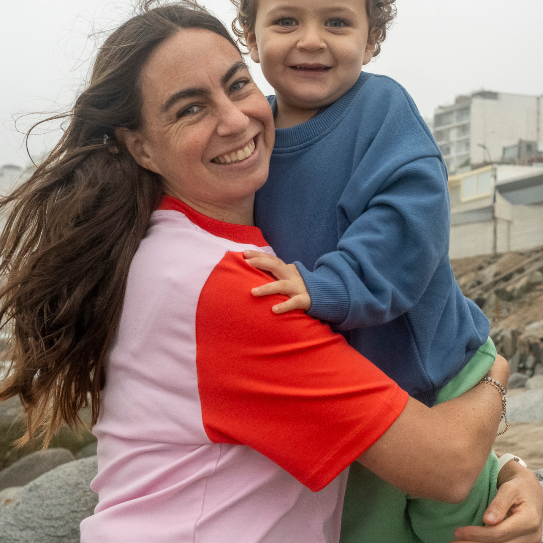 Pink with Engine Red | Woman holding a child on a rocky beach with buildings in the background