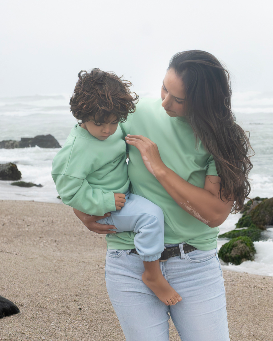 Grass Green | Woman holding a child on a beach with ocean waves in the background