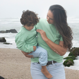 Grass Green | Woman holding a child on a beach with ocean waves in the background
