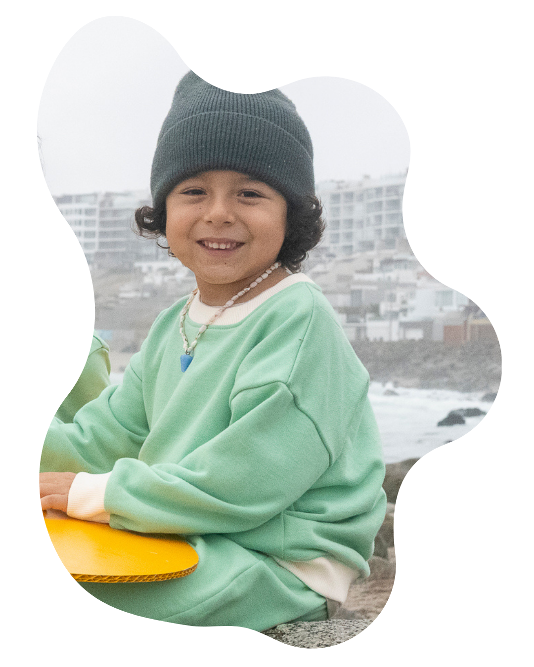 Grass Green | Child wearing a green sweater and gray beanie holding a yellow frisbee with a blurred cityscape background