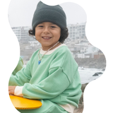 Grass Green | Child wearing a green sweater and gray beanie holding a yellow frisbee with a blurred cityscape background