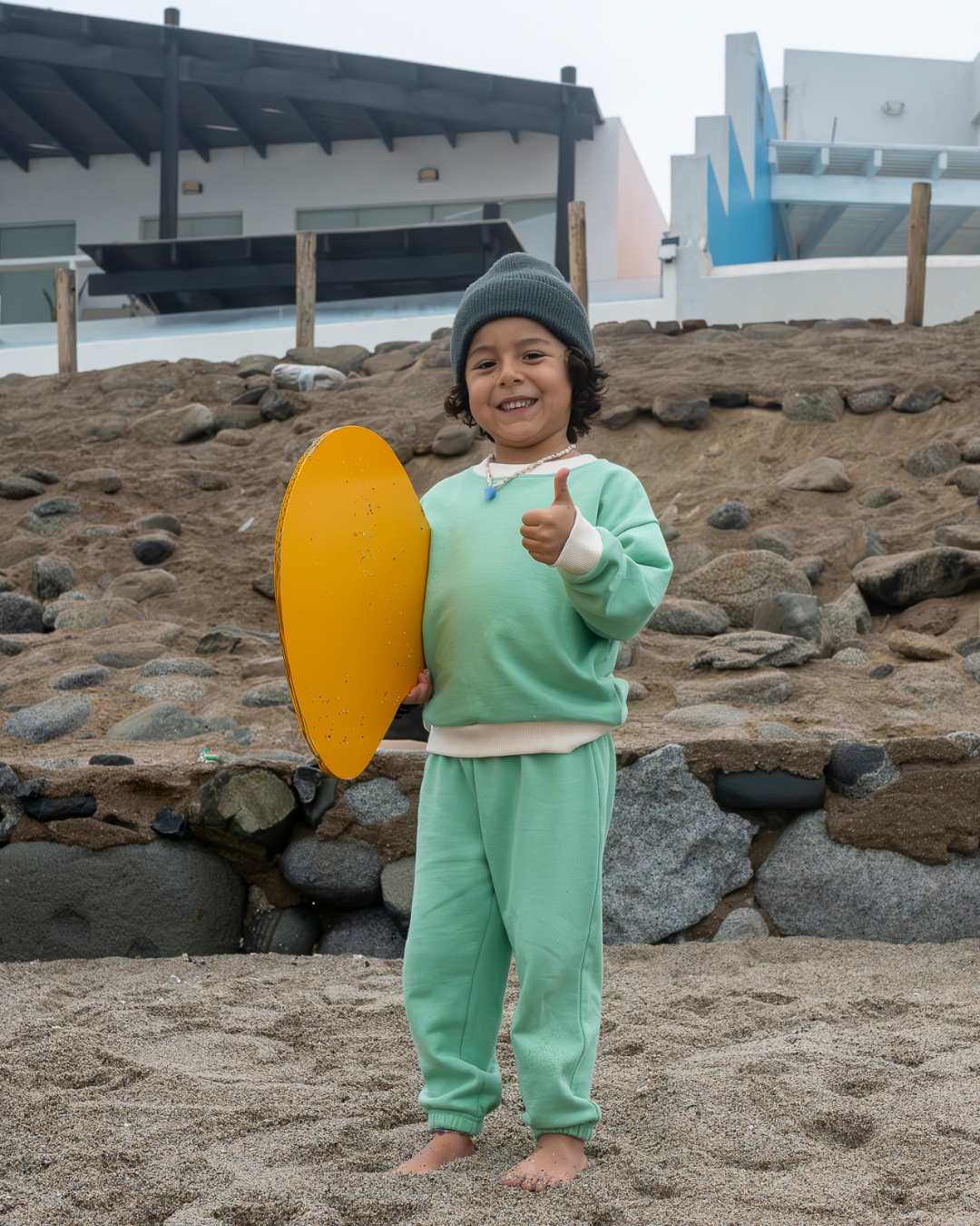 Grass Green | Child in green outfit holding a yellow object on a rocky beach.