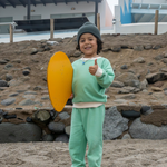 Grass Green | Child in green outfit holding a yellow object on a rocky beach.