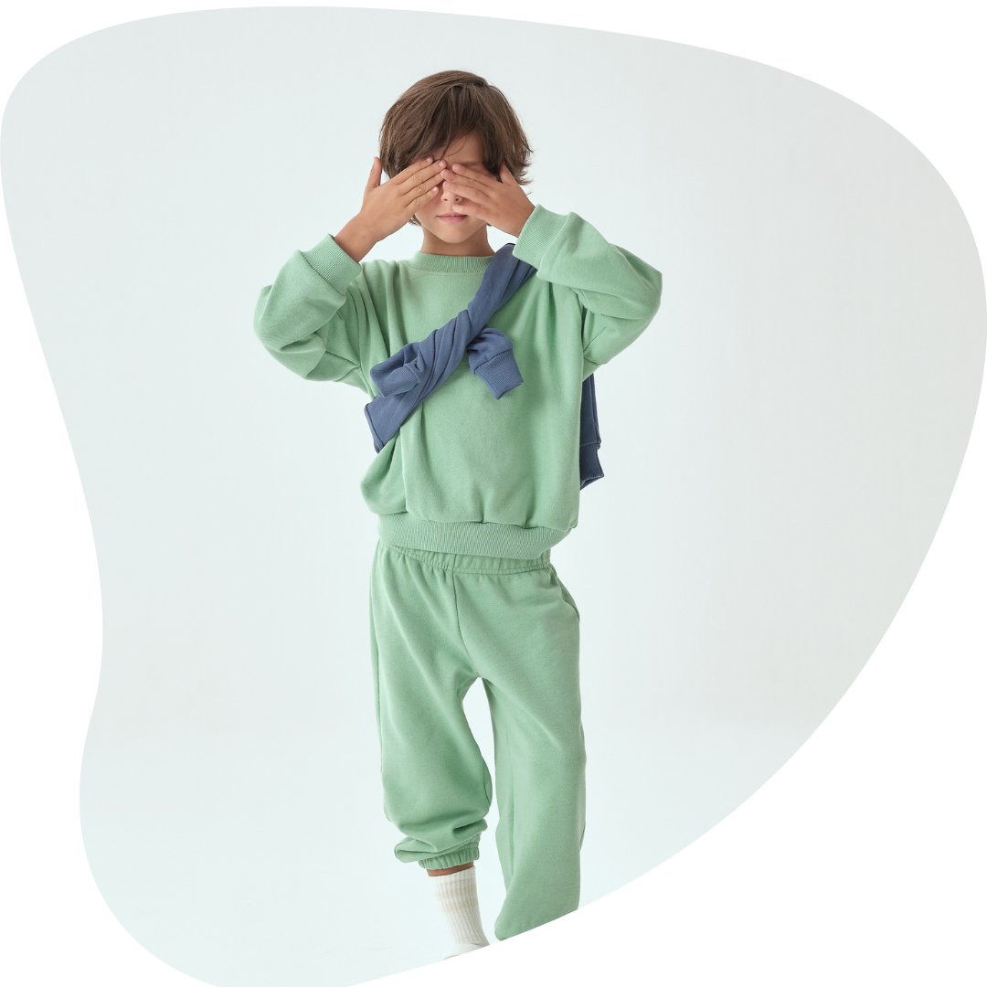 Grass Green | Child wearing a light green outfit with a blue sweater over shoulders on a white background