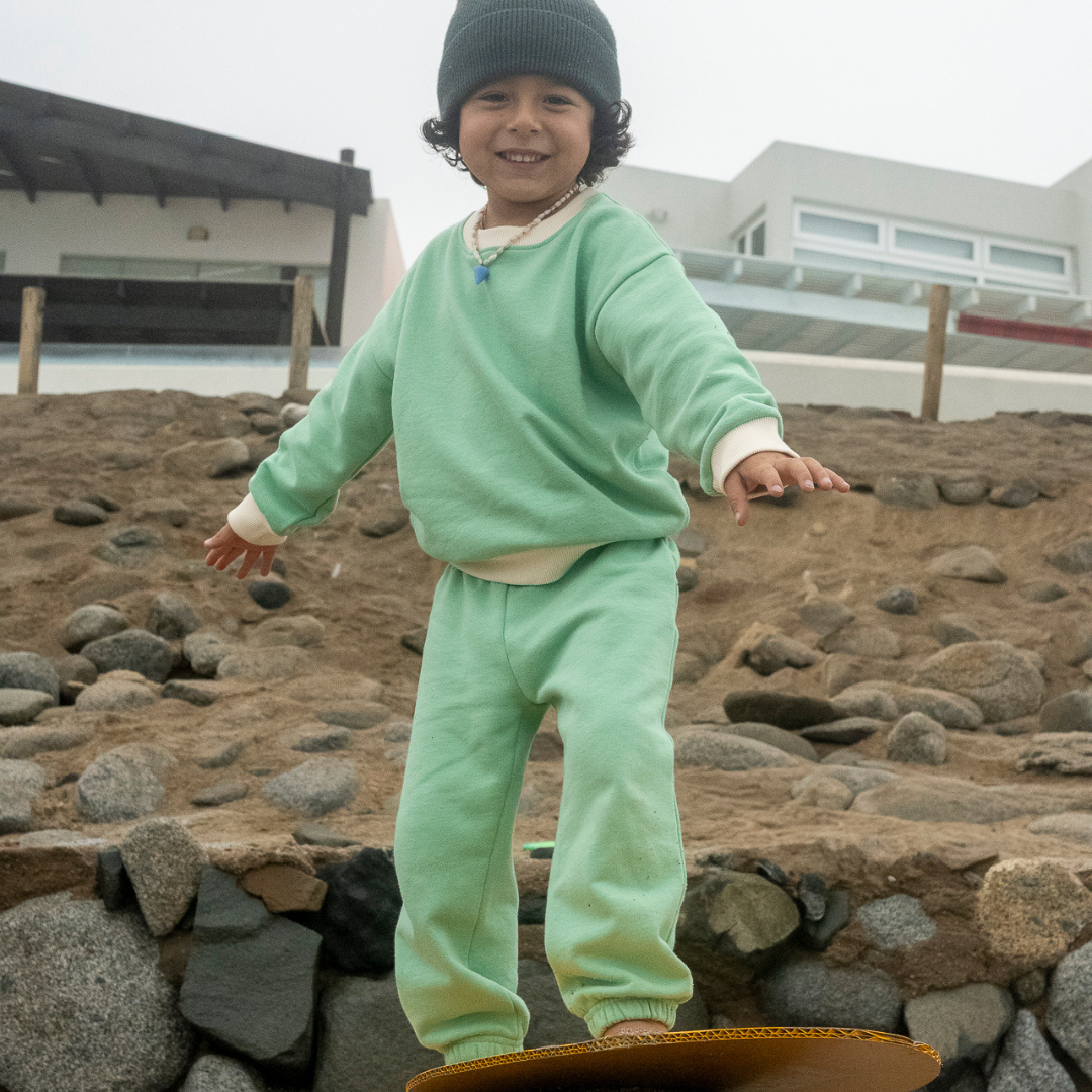 Grass Green with White | Child in green outfit standing on a skateboard by a stone wall.