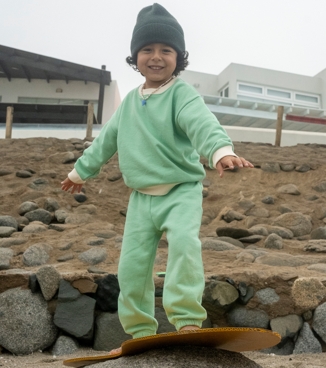 Grass Green with White | Child in green outfit standing on a skateboard by a stone wall.
