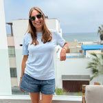 Light Blue | Woman wearing sunglasses and a blue t-shirt with denim shorts standing by a window with a view of the ocean.